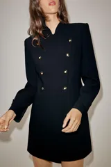 Vestido tipo blazer de corte estructurado, color negro, con cuello subido y cierre frontal cruzado con botones metálicos dorados. Presenta mangas largas con trabillas en los puños y bolsillos de vivo decorativos en el frente.