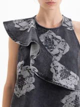 Vestido corto de denim con lavado gris y estampado floral en contraste. Presenta un diseño sin mangas con escote cerrado y un volado asimétrico en el frente que aporta movimiento. Posee un calce suelto y cierre en la espalda.