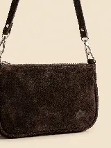 Cartera pequeña tipo bandolera confeccionada en gamuza color marrón oscuro, con cierre superior metálico y correa ajustable con herrajes plateados. Presenta un detalle de logo grabado en la parte inferior.