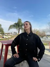 Sobre camisa negra de corte recto, con cuello clásico, cierre frontal con botones dorados, bolsillos de parche en el pecho y detalle de calados en el ruedo.