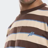 Remera de manga larga con diseño de rayas horizontales en tonos marrón, beige y azul. Presenta un corte boxy holgado y un efecto de superposición en las mangas con puños en color marrón sólido. Incluye un logo bordado en el pecho y en la manga derecha.