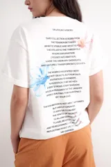 Remera de corte oversize, color blanco, con cuello redondo y mangas cortas. Presenta una estampa frontal con el texto 'ATELIER (UNVEILING VISIONS)' y tres recuadros con ilustraciones de flores.