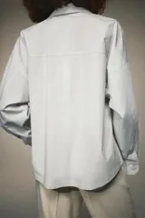 Camisa de popelín color blanco, con cuello solapa, escote en pico y cierre frontal con botones. Presenta un corte holgado y mangas largas con puños rectos.