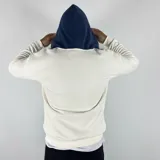 Canguro blanco hueso de algodón con felpa, capucha forrada en azul marino, cierre parcial con cremallera, cordón ajustable y bolsillo tipo canguro con solapa.