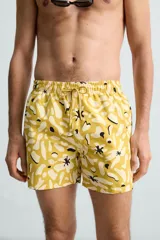 Bañador de hombre de tejido técnico ripstop, color mostaza con estampado floral abstracto en blanco y negro. Presenta cintura elástica ajustable con cordón, bolsillos laterales y bolsillo trasero de plastrón.