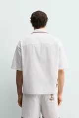 Camisa de corte relajado confeccionada en algodón, con cuello bowling y manga corta. Presenta un bolsillo en el pecho con vivo a contraste y bordados decorativos en el frente. Cierre frontal mediante botones.