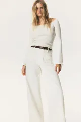 Top corto de punto acanalado color blanco, con escote amplio que deja los hombros al descubierto y leve escote en V. Mangas largas acampanadas con ribete de volados en puños y bajo.