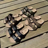 Sandalia tipo Birkenstock con doble tira ajustable de cuero sintético negro y hebillas metálicas. Cuenta con plantilla anatómica de corcho y suela de goma.