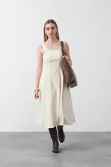 Vestido midi sin mangas, de color crema, con escote recto y silueta evasé. Presenta costuras a contratono que recorren el frente y la falda, creando un diseño estructurado y estilizado.