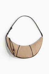 Bolso de hombro con forma de media luna, cuerpo principal en tela color beige y ribetes y correa en cuero sintético negro. Presenta cierre superior con tiradores largos de cordón negro anudado y argollas metálicas doradas.