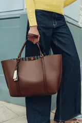 Bolso tipo shopper de color marrón, con diseño minimalista, doble asa de hombro y cierre magnético. Incluye un colgante decorativo con detalle de cinta en tonos claros y un bolsillo interior amplio.