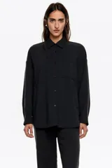 Camisa de corte oversize color negro, confeccionada en mezcla de algodón y lyocell con textura. Presenta cuello camisero, cierre frontal con botones, manga larga con puños abotonados y un bolsillo de plastrón en el pecho.