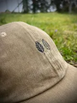 Gorra tipo dad hat en color gris lavado, con diseño de dos hojas bordadas en hilo negro en la parte frontal. Cuenta con visera curva y estructura flexible.