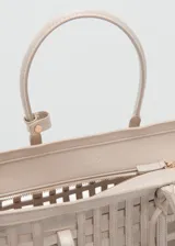 Bolso cesta de diseño trenzado en color beige, con dos asas cortas de mano y un asa larga ajustable y desmontable. Incluye detalle de charm colgante y herrajes metálicos.
