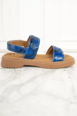 Sandalias con estampado de piel de serpiente en tonos azules, con dos tiras anchas sobre el empeine y una tira alrededor del tobillo. La suela es plana y de color marrón claro.