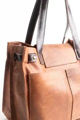 Cartera tipo tote de cuero color beige con diseño de flecos en la solapa frontal. Cuenta con manijas de cuero oscuro, cierre metálico tipo torniquete y detalles de herrajes dorados.