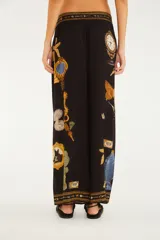 Pantalón largo de silueta amplia, color negro, con diseño estampado de diversos objetos como relojes, palmeras, animales y marcos estilo barroco. Cuenta con cintura elastizada y detalles decorativos en los bordes de la cintura y los tobillos.