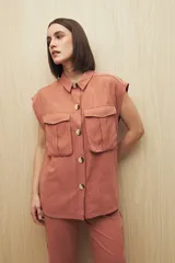 Chaleco color beige con cuello camisero, sin mangas, cierre frontal con botones y cinturón de lazo. Presenta bolsillos de parche en el pecho y pinzas verticales en el frente.