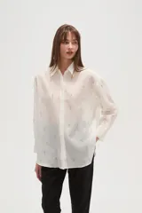 Camisa blanca de poliéster, de corte ancho y manga larga, con cuello redondo y apliques de strass y tachas distribuidos por toda la prenda.