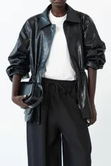 Chaqueta semi-larga negra con acabado brillante o efecto charol, cuello camisero y frunce ajustable en la cintura. Presenta cierre frontal y bolsillos prácticos.