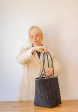 Cartera tipo tote de gamuza color marrón, con correas trenzadas y detalle de piedras naturales incrustadas en líneas verticales. Cuenta con cierre metálico y forro interior de tela.