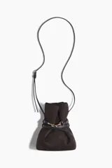 Cartera tipo bombonera de color marrón oscuro, confeccionada en tejido revestido con acabado mate. Presenta un diseño fruncido en la parte superior ajustado por una tira delgada con cierre de mosquetón metálico dorado. Incluye una correa larga y fina ajustable para llevar cruzada.