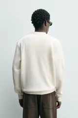 Camiseta relaxed fit color crema, con cuello redondo tipo panadero y cierre frontal de tres botones. Manga larga y acabados en rib.