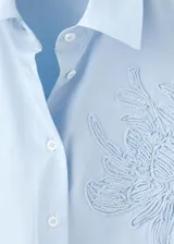 Camisa celeste de corte oversize, con mangas largas, cuello camisero, cierre frontal con botones y detalle de bordado floral en el pecho.