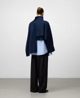 Cazadora corta de gabardina azul marino con cuello chimenea y cierre frontal cruzado con botones. Presenta un diseño asimétrico con una pieza de tejido integrada en la manga para llevar a modo de chal.