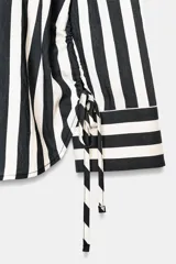 Camisa de cuello solapa con escote pico y manga larga. Presenta un bolsillo delantero de plastrón y bajo con aberturas laterales con cordones ajustables. Cierre frontal con botones.