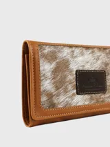 Billetera de cuero combinada en tonos camel y marrón, de formato rectangular con solapa. Cuenta con un parche central decorativo con logo grabado y detalles de costura perimetral.