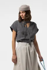 Camisa gris de corte relajado, con escote en V y lazo largo incorporado en el cuello. Mangas cortas con vuelta y frunce sutil en los hombros.