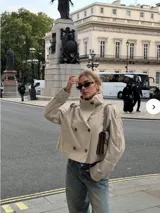 Chaqueta corta tipo trench color beige, con doble botonadura frontal, cuello alto y mangas largas con puños ajustables. Presenta un diseño estructurado con hombreras y solapas.