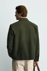 Sobrecamisa negra de corte relajado, confeccionada en tejido con mezcla de algodón elástico. Presenta cuello solapa, cierre frontal de cremallera, bolsillos de plastrón en el pecho y bolsillos de vivo en la cadera.