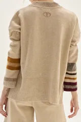 Sweater tejido de lambswool color beige, con cuello redondo y puños acampanados con rayas de colores (marrón, mostaza, bordó y beige). Incluye un detalle de temporada hecho a mano en la espalda.