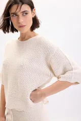 Sweater color crudo de manga corta, confeccionado en tejido de punto con diseño de textura fantasía, cuello redondo y corte holgado.