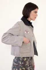 Campera corta de jean color visón, con cuello desmontable de tela engomada, cierre frontal y bolsillos plaqué con solapa en el pecho. Cuenta con un parche bordado en el delantero.