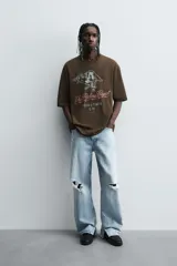 Camiseta de algodón color marrón oscuro, corte relajado (relaxed fit) y manga corta. Presenta un estampado frontal con un lobo o cánido corriendo, el texto "The Riders Project", "VISION & TEMPER" y "0.79 per gallon" en tonos grises y rosados.