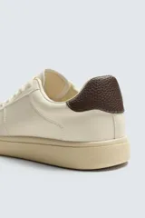 Zapatilla deportiva de diseño minimalista en color crema, con detalle de contraste en color marrón oscuro en el talón y suela de goma con volumen.