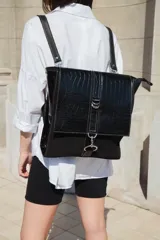 Mochila negra de cuero vacuno con textura tipo cocodrilo en la solapa superior y detalles de costuras blancas. Tiene mosquetón de seguridad exterior, cierre interior, dos bolsillos interiores (uno con cierre y otro para celular), sujetador de llaves, forro completo y tiras ajustables. Puede usarse como mochila o como cartera.