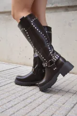 Bota de caña media en cuero sintético negro, con suela track, cierre lateral y detalle de tachas metálicas en la parte superior y en una tira decorativa sobre el tobillo.