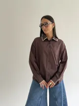Camisa oversize de color marrón con cuello camisero y puños con detalle de bordado festoneado en color blanco. Presenta cierre frontal de botones y mangas largas.
