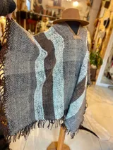 Poncho de lana virgen tejido en telar con diseño geométrico a rayas en tonos gris, marrón y celeste, con apliques de cuero en el escote y flecos en el ruedo.