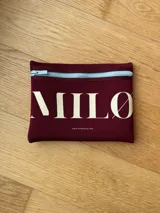 Clutch o neceser de neopreno color bordó con cierre de cremallera en la parte superior. Estampado frontal con la palabra "MILØ" en color crema y la dirección web "WWW.STOREMILO.COM" debajo.