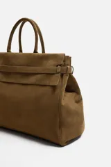 Bolso de mano extragrande color marrón oscuro, estilo tote o satchel, fabricado en piel. Presenta dos asas de mano y un detalle de cinta ajustable con cierre de solapa en la parte frontal superior.