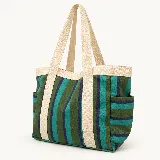 Bolso tipo tote bag de lona con diseño de rayas verticales en tonos verde, turquesa y azul marino. Cuenta con asas anchas de hombro en color beige y bolsillos laterales. Incluye un parche circular con el logo de la marca en el frente.