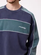 Sweater de cuello redondo marca Rusty, con diseño de bloques de color en azul marino y verde, con detalles de líneas blancas. Presenta el logo de la marca bordado en el pecho.