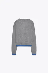 Chaqueta de punto gris de manga larga, cuello redondo y cierre frontal con botones oscuros. Presenta ribetes y detalles en color azul eléctrico en el frente, puños y ruedo.