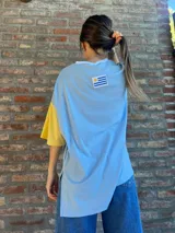 Remera de corte oversize confeccionada en tejido de punto acanalado color celeste, con mangas cortas en color amarillo y detalle de parche bordado con la bandera de Uruguay en la parte superior de la espalda.
