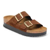 Sandalia de dos correas con hebillas metálicas doradas, confeccionada en gamuza color marrón. Cuenta con plantilla anatómica de corcho y suela de plataforma dentada en color negro.